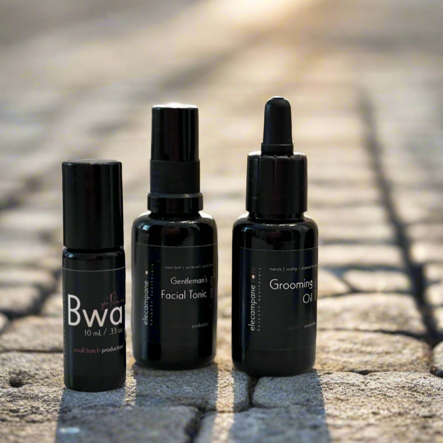 Three black skincare bottles labeled 'Bwa', 'Facial Tonic', and 'Grooming Oil' on a stone surface.