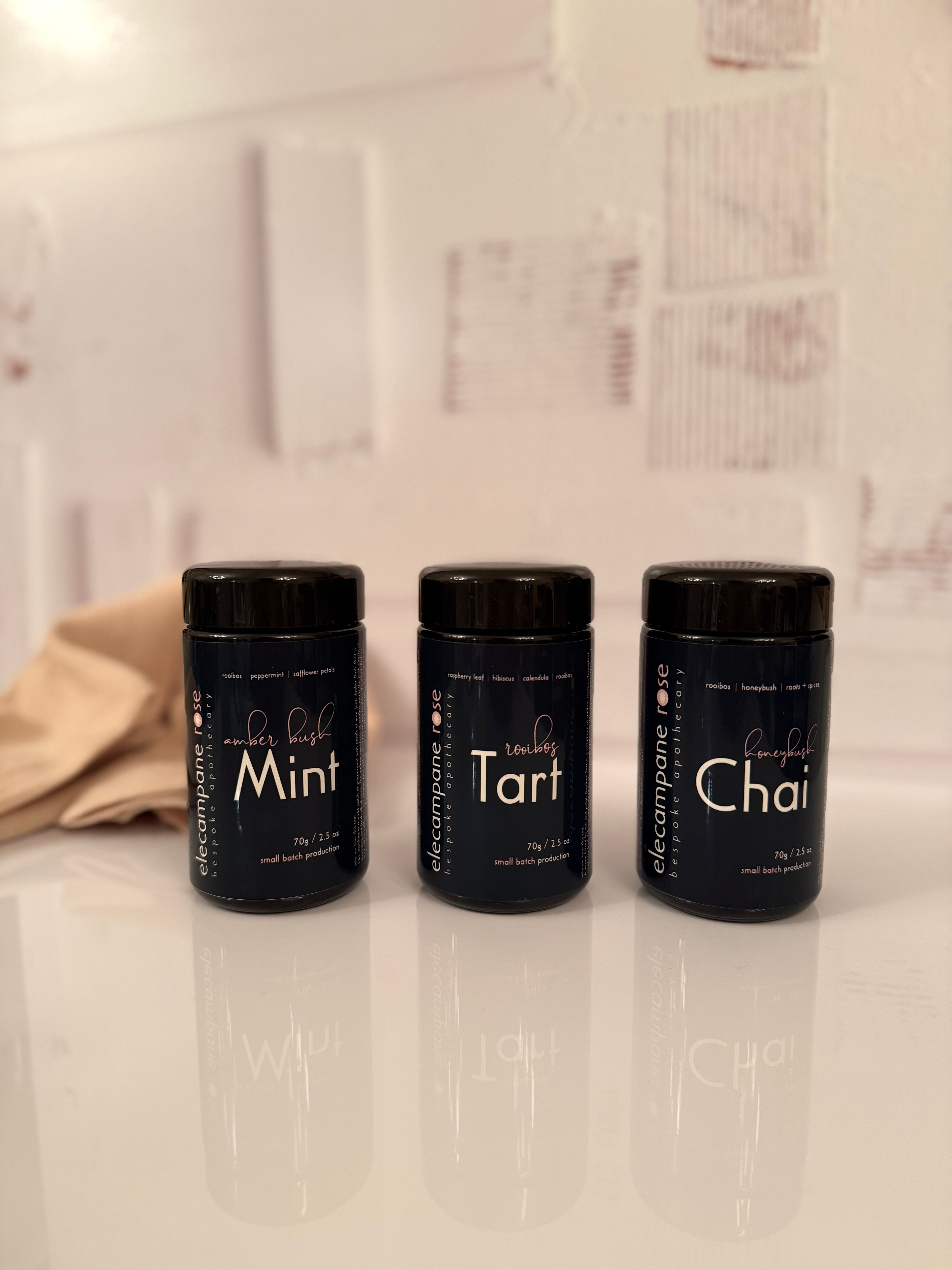 Three black containers labeled 'Mint', 'Tart', and 'Chai' on a white surface with a tiled wall background.